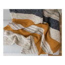 A chunky knit throw blanket with horizontal stripes in cream, mustard yellow, and dark charcoal gray, draped over a light-colored sofa.