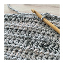 Close-up of a crocheted fabric swatch in heather gray, white, and soft beige yarn, with a wooden crochet hook inserted into the stitches on a light wood surface.