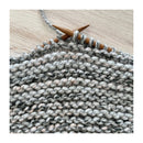 Close-up of a knitted fabric swatch in a variegated gray, white, and soft pink yarn, held on wooden knitting needles over a light wood background.