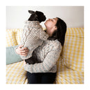 A woman in a chunky knit beige sweater holds a small black dog wearing a matching cable-knit sweater while sitting on a yellow-and-white checkered couch.