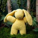 Yellow crochet bunny toy in a natural setting with plants and grass.