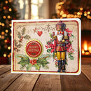A rectangular Christmas greeting card with a nutcracker illustration, holly leaves, red berries, and a round 'Season's Greetings' badge, displayed on a wooden surface with blurred holiday lights in the background.