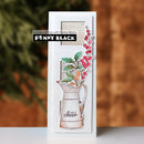 A handmade greeting card featuring a white cardstock base with a layered die-cut design of a vintage metal pitcher filled with green leaves and red berries, with a black and white 'PENNY BLACK' brand sticker on the upper left. The card is photographed standing upright on a white surface against a blurred brown and green background.