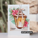 A handmade greeting card featuring a lantern with a candle, pink poinsettias, and green leaves, photographed standing upright against a softly blurred outdoor background. The card includes the text 'Christmas in full bloom' and a visible 'Penny Black' brand mark at the bottom.