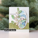 A handmade greeting card featuring a winter illustration of a child in a blue hat and green scarf holding a mug, with holly leaves and the words 'peace & goodwill'. The card is displayed upright against a blurred green holiday tree background, with a 'Penny Black' brand label at the bottom.