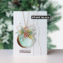 A handmade Christmas greeting card by Penny Black, featuring a watercolor ornament design with holly leaves and a pinecone, on a white card with textured paper details and a blurred green holiday background.