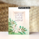 A handmade greeting card by Penny Black featuring a watercolor illustration of green leaves and white berries with a warm message for Christmas. The card is displayed upright on a white surface against a soft, blurred brown background.