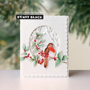 A handmade greeting card by Penny Black featuring a white scalloped border and a die-cut oval design with a bird, holly leaves, and red berries. The card is displayed on a light surface with a blurred green pine background.