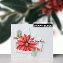 A handmade greeting card featuring a layered red poinsettia flower with green holly leaves and a small gold center, displayed against a blurred holiday background. The card has a white embossed border and a black banner with the brand name 'PENNY BLACK'.