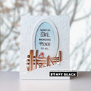 A handmade greeting card featuring a white embossed snowflake background, a central oval with a light blue center and the quote 'BORN IN LOVE, BRINGING PEACE TO ALL', a brown wooden fence, and a small red bird. The card is displayed on a white surface with a blurred winter tree background.