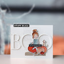 A handmade layered card by Penny Black featuring a cute cartoon girl sitting on orange pumpkins with a small cat, set against a large white 3D 'BOO' lettering. The card has a black label reading 'papercrafting with Penny Black' and 'PENNY BLACK' at the top, with a soft, blurred background.