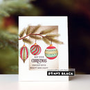 A handmade Christmas greeting card featuring a watercolor pine branch with three striped ornaments in red, green, and gold, with the text 'MAY YOUR CHRISTMAS UNFOLD WITH BEAUTY AND LIGHT.' The card is displayed on a white surface with a blurred festive background, and a 'papercrafting with Penny Black PENNY BLACK' brand sticker is visible at the bottom.