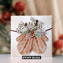 A handmade Christmas greeting card featuring layered paper cutouts of knit mittens, holly leaves, pinecones, and a banner reading 'simple joys of Christmas.' The card is photographed upright against a softly blurred holiday background with red berries and greenery.