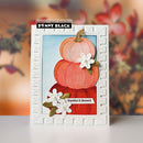 A handmade autumn-themed greeting card by Penny Black, featuring three layered watercolor pumpkins in shades of orange, red, and peach, accented with white paper flowers and green leaves. The card is set against a white embossed border and displayed on a clean white surface with a blurred warm-toned background.