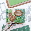 A handmade layered card featuring pinecones and green pine needles on a wood-grain and green patterned background, with a scalloped oval label reading 'peace ON EARTH.' Silver die-cut embellishments and patterned paper sheets are arranged around it on a white surface.
