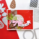A handmade Christmas greeting card with a red background, decorated with a pink bow, pinecones, green pine needles, white snowflakes, and a white banner reading 'Joy to the world'. The card is styled on a white surface with craft dies and other cards around it.