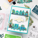 A handmade layered card featuring a teal and white holiday cityscape with houses, a snow globe, a reindeer, pine trees, and gift boxes, layered over a white cardstock base. The card is styled on a flat-lay desk with craft supplies, including an ink pad, a stamp sheet, and other holiday-themed items.
