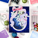 A handmade layered card featuring a dark blue background with vertical stripes, a large pink and lavender ribbon bow, white leafy branches, and small pink berries. The card is styled in a flat-lay with craft supplies, stamps, and ink pads around it.