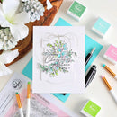 A handmade greeting card with a layered white background and a clear stamp design reading 'Sending Christmas Cheer' with blue flowers and green leaves, styled in a flat lay with crafting supplies and flowers.