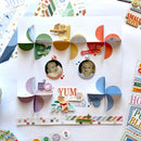 A colorful scrapbook layout card featuring three paper pinwheel overlays, circular photo cutouts of babies, decorative text elements like 'YUM is your name,' and coordinating paper strips and stickers, arranged on a white surface with other craft supplies around it.