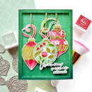 A handmade Christmas ornament card set displayed in a green presentation box, featuring three colorful ornament cutouts with floral and leaf patterns, gold hanging strings, and a die-cut sentiment reading 'Warm wishes this Christmas.' Surrounding props include stamp pads, embossed metal ornaments, and a makeup brush on a white background.