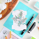 A handmade greeting card with layered white paper tabs, a floral die-cut design in teal and green, and the phrase “Season’s Greetings,” styled on a turquoise sheet with pens and craft supplies around it.