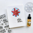 A handmade greeting card with a circular cutout showing a red poinsettia flower, placed on a white surface with craft supplies including a metal die, clear stamped phrases, and a small bottle of gold liquid.