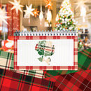 A spiral-bound notepad with a red plaid border and a white center panel featuring a green and red rose illustration, styled on top of red plaid fabric with a blurred Christmas tree and hanging star decorations in the background.