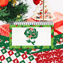 A spiral-bound notepad with a green Christmas-themed border and a white lined center page featuring a green rose illustration, styled on top of red and green holiday gift-wrapped paper and near a wooden Santa decoration.