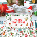 A spiral-bound Christmas greeting card or notebook with 'MERRY CHRISTMAS' in red text, surrounded by festive illustrations and placed on top of other holiday cards. The scene is styled with wrapped gifts, a knit sweater, pinecones, and a blurred Christmas tree background.