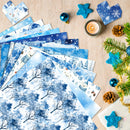 A styled flat-lay of blue winter-themed patterned paper sheets fanned out on a light wood surface, surrounded by holiday decorations including pine branches, blue star ornaments, a lit candle, and small gift boxes.