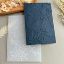 A dark navy embossed card or paper sheet with a raised bird and floral branch design, placed over a translucent white embossed sheet on a neutral surface with dried floral props.