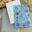 A floral embossed card or art print with blue and purple flowers on a light blue background, shown alongside a white embossed sheet and a translucent embossed template. The scene is styled with dried botanicals on a neutral surface.