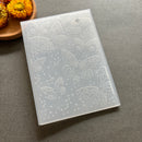 A translucent plastic stamping plate with embossed butterfly designs and small bubble patterns, shown on a gray surface with dried flowers in the corner.