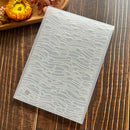 A translucent silicone mold with a repeating wave and fish pattern, displayed diagonally on a dark wood background with dried flowers in the corner.