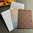 Three textured embossed paper sheets with a woven basketweave pattern are displayed in different colors: white, iridescent gold-to-blue, and brown. A translucent sheet is visible in the background on a dark gray surface.