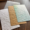 Three embossed botanical paper sheets with floral relief patterns in white, tan, and sage green, arranged on a dark wood surface.