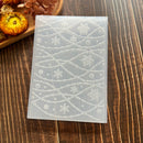 A translucent silicone stamping or embossing plate featuring a white snowflake and wavy line pattern, displayed on a dark wood background with dried flowers in the corner.