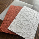 Two embossed patterned cards or paper sheets with snowflake and dotted line motifs, one in terracotta red and one in white, shown on a dark wood surface with a translucent template or sheet partially visible behind them.