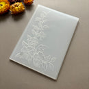 A translucent white acrylic or plastic plaque with an embossed floral vine design, photographed on a neutral taupe background with dried orange flowers in the corner.