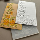 Three rectangular cards or sheets stacked on a neutral surface: a tan card with raised yellow flowers and green leaves, a white embossed floral card, and a translucent gray sheet showing an embossed floral design.