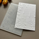A white embossed greeting card with a winter holiday design featuring two candles, pine branches, and snow-like dots, shown alongside a translucent silicone stamp mold on a neutral gray background.