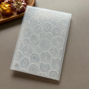 A translucent silicone mold with a raised circular tree-ring pattern, shown at an angle on a neutral gray surface with dried flowers in the background.