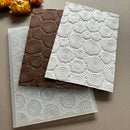 Three embossed paper sheets with circular rose patterns in brown, white, and translucent white, arranged on a neutral gray background with dried flowers in the top left corner.