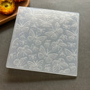 A square translucent silicone mold with embossed butterfly and floral patterns, shown on a gray surface with a dried flower prop in the top-left corner.