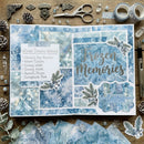 A styled flat-lay of a scrapbook journaling page with winter-themed blue and white patterned paper, silver glitter text reading 'Frozen Memories,' decorative botanical stickers, and surrounding crafting supplies on a wooden surface.