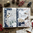 An open handmade scrapbook journal displayed on a rustic wooden table, surrounded by winter-themed decorative props like pinecones, red berries, snowflakes, and a vintage camera. The left page features a chickadee and the words 'Winter Serenity' with snowy branches and evergreen trees.