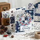 A handmade winter-themed greeting card displayed on a rustic white wooden table, surrounded by crafting supplies, pinecones, twine, and other patterned cards. The card features a patchwork design of navy blue and white floral squares, two birds perched on a branch with red berries in a wreath, and the text 'WINTER Serenity'.