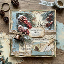 A square handmade junk journal with a rustic winter theme, decorated with pinecones, evergreen branches, musical notes, vintage-style stamps, and a small copper bell, tied with twine on a wooden surface.