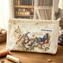 A cream-colored quilted cosmetic pouch with embroidered butterflies, flowers, and a 'AGOGO ORIGINAL' label, displayed on a wooden table with sewing supplies in a cozy room setting.